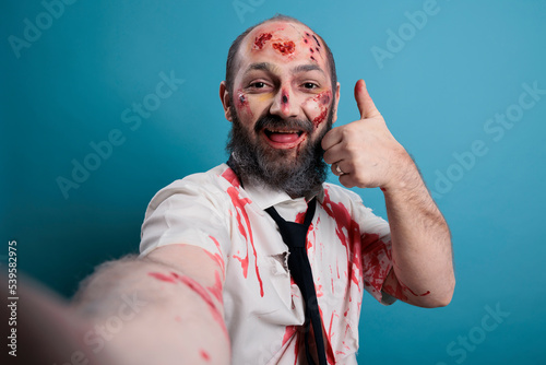 Apocalyptic monster doing thumbs up sign in front of camera, taking picture on smartphone. Creepy evil brain eating corpse showing like and approval symbol, scary horror zombie.