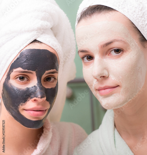 Two smiling young women in bathrobes and towels putting facial mask on face after shower while sitting in bedroom bed