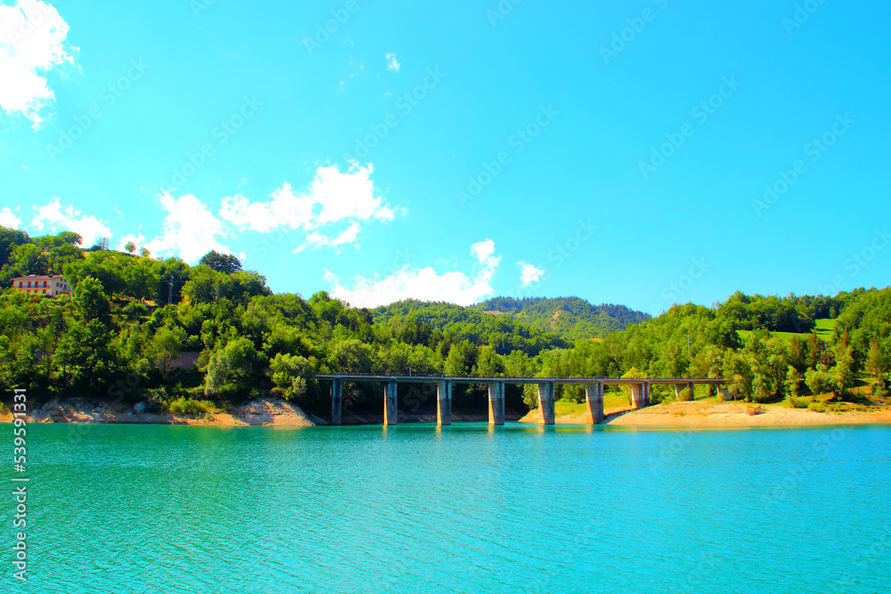 Tidy view of Gerosa Lake with a massive bridge suspended above the pure slightly rippled blue waters and reaching two hills chock full of vegetation and some buildings under a clear blue sky