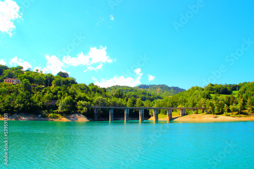 Tidy view of Gerosa Lake with a massive bridge suspended above the pure slightly rippled blue waters and reaching two hills chock full of vegetation and some buildings under a clear blue sky