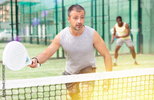 Young adult man playing paddle tennis doubles match with male partner at warm sunny day © JackF