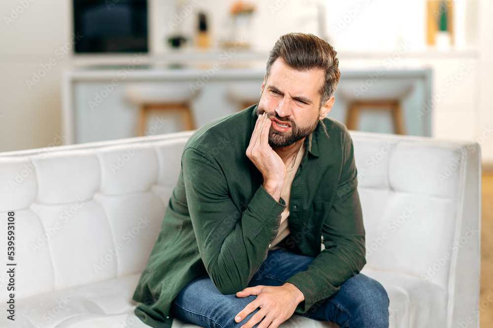 Toothache. Unhappy upset caucasian sad man sits on the couch at home, holds his hand near his cheek, grimaces, has an acute toothache, needs a consultation with a dentist doctor Toothache. Unhappy upset caucasian sad man sits on the couch at home, holds his hand near his cheek, grimaces, has an acute toothache, needs a consultation with a dentist doctor