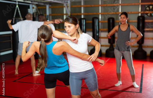 Latin woman training elbow strike with another woman during group self-protection training. Asian woman instructor standing nearby and correcting them.