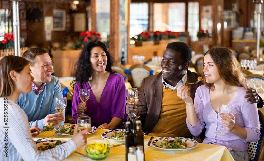 Multiethnic group of positive men and women sitting at table in restaurant, laughing and drinking wine.