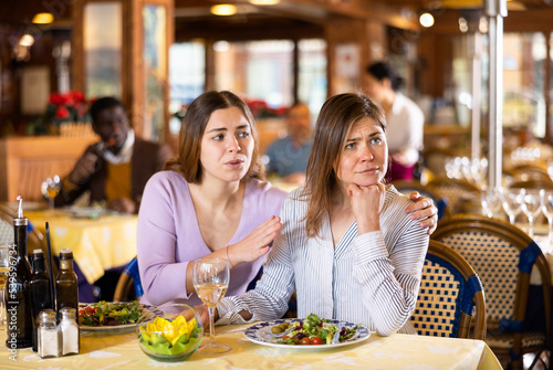 Caring woman calming her upset female friend while sitting together at table in cozy restaurant during dinner..