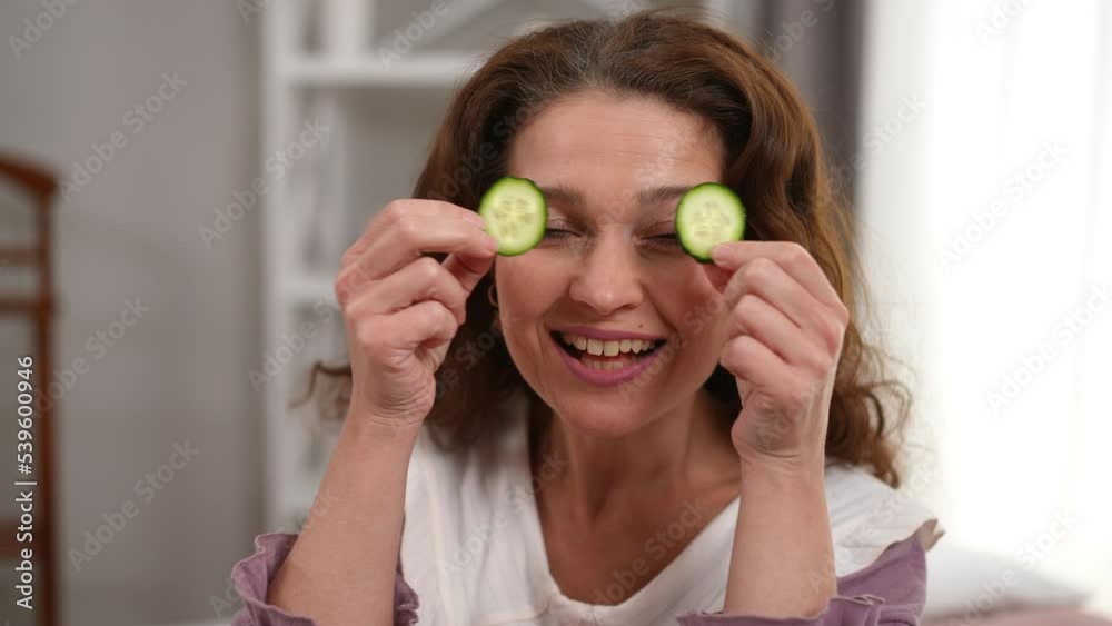 Close-up portrait of joyful Caucasian mature woman having fun doing beauty routine at home on weekend. Front view of happy cheerful lady looking at camera covering eyes with cucumber slices smiling Close-up portrait of joyful Caucasian mature woman having fun doing beauty routine at home on weekend. Front view of happy cheerful lady looking at camera covering eyes with cucumber slices smiling