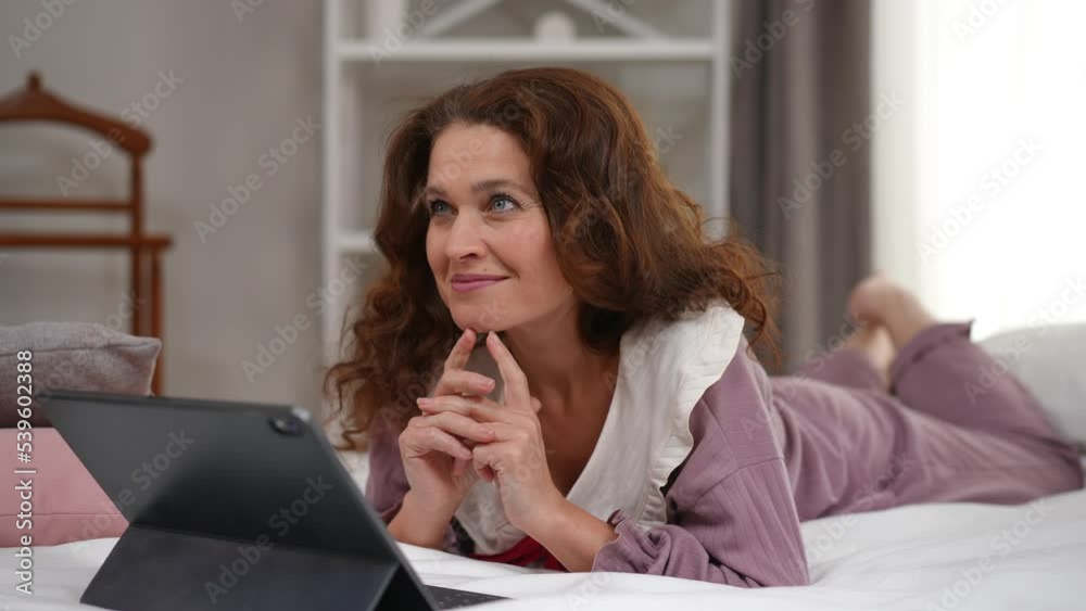 Smiling happy woman watching romantic movie on digital tablet dreaming lying on bed in the morning. Portrait of charming mature Caucasian lady in pajamas enjoying leisure at home Smiling happy woman watching romantic movie on digital tablet dreaming lying on bed in the morning. Portrait of charming mature Caucasian lady in pajamas enjoying leisure at home