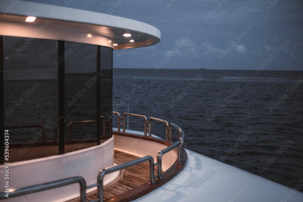 Fototapeta premium A low-key view of a part of a safari deck next to the bow of the boat or of its stern, with a passage fenced with a chromed fencing and a mirrored glass windows of a cockpit; a seascape behind it
