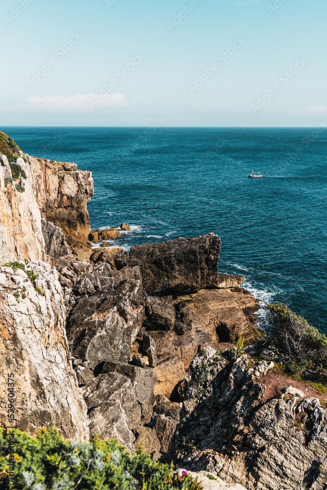 A vertical scenery of a rocky cliff with sparse vegetation, plenty of huge stones, and a lonely boat behind the shoreline floating on turquoise waves of the ocean with a horizon in the background