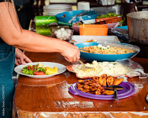 Mulher se servindo comida típica Brasileira, mesa de madeira, com pratos brancos potes azuis, almoço familiar 