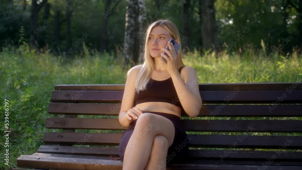 Attractive young woman talking on phone in park. blonde in black sports top sits on bench and chats on smartphone. girl is glad to hear answer and smile. Attractive young woman talking on phone in park. blonde in black sports top sits on bench and chats on smartphone. girl is glad to hear answer and smile.