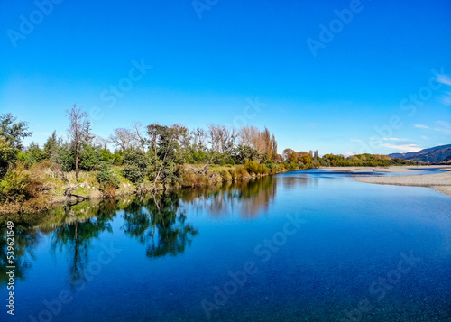 A landscape image of trees reflecting in the still blue waters of the Wairau River on the Marlborough Region of the South Island of New Zealand
