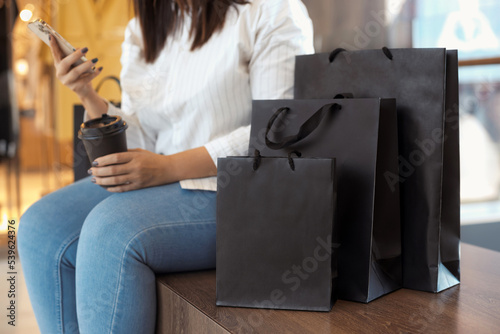 Mock up paper bags with faceless woman on background relaxing after shopping, holding mobile phone and cup of coffee. Black friday concept. Mockup and copy space. 