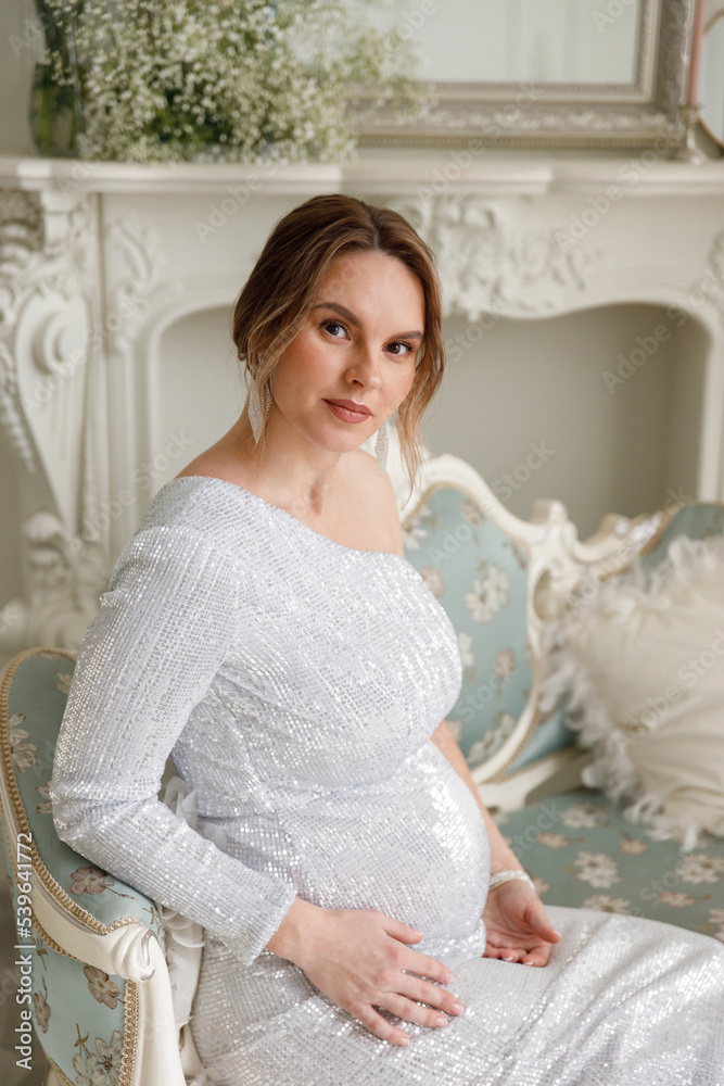 beautiful pregnant woman in evening dress sits on a sofa in a white room. The concept of motherhood and the birth of children. Glamor and beautiful life