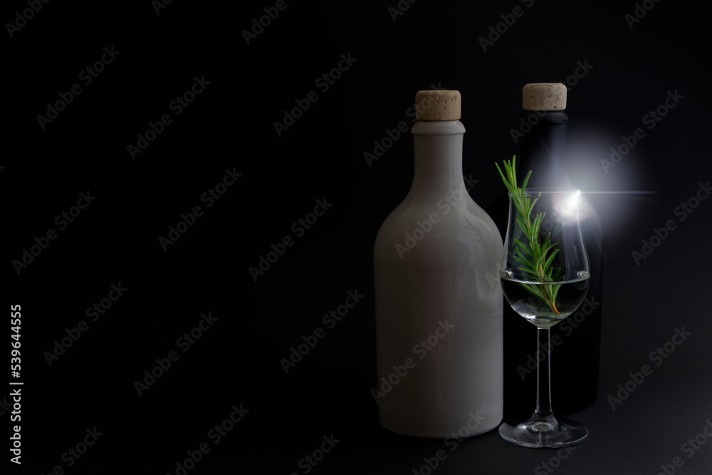 White and black ceramic gin bottles with beautiful glass of gin and green rosemary on black background and interesting light effect. Low light photo shot. Copy space for your design. Web Banner. White and black ceramic gin bottles with beautiful glass of gin and green rosemary on black background and interesting light effect. Low light photo shot. Copy space for your design. Web Banner.