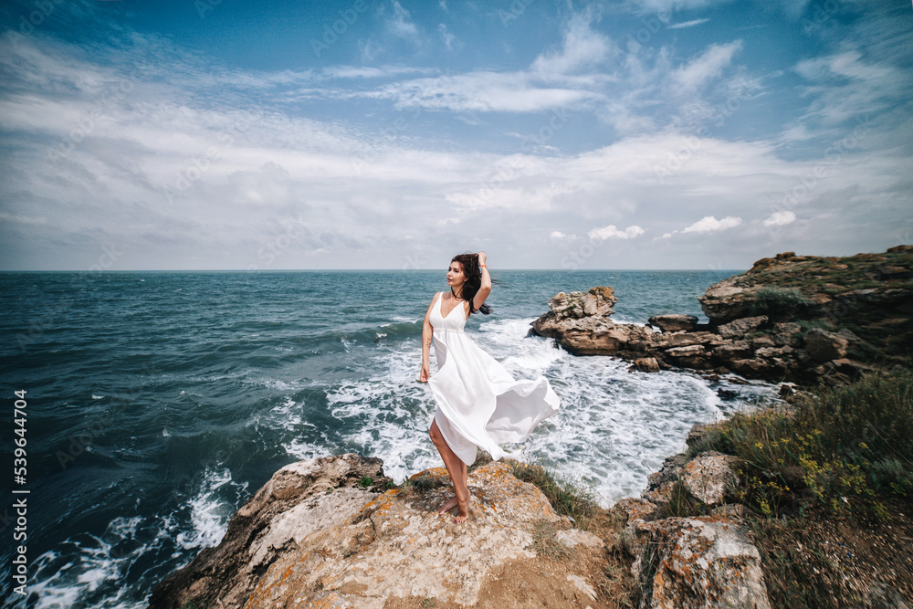 A beautiful young woman stands on the rocks in a white light dress against the backdrop of the sea.
