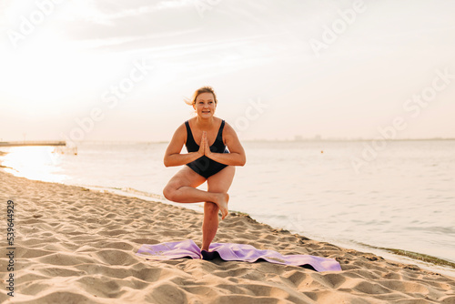 Fototapeta Naklejka Na Ścianę i Meble -  Caucasian woman practicing yoga at seashore sandy beach on sunrise. Womens health and wellness. Sports body positive. Real instructor poses