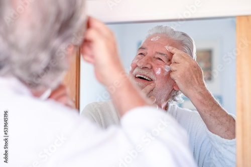 Skin care. Handsome senior man applying cream at his face and looking at himself with smile while standing in front of the mirror