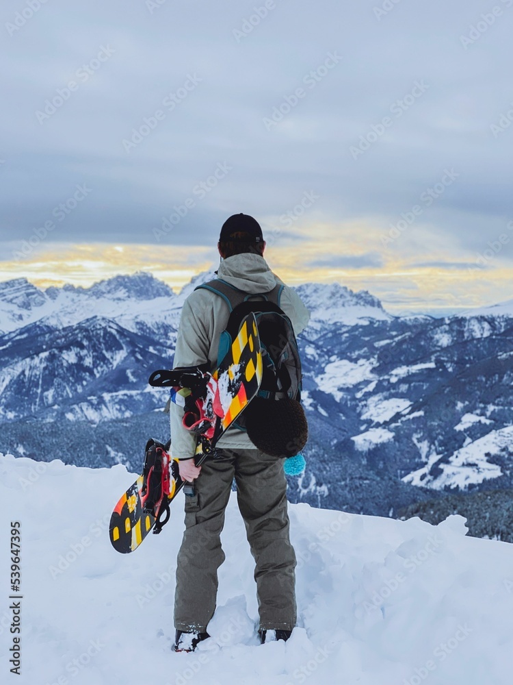 Naklejka premium Vertical shot of a snowboarder looking at the snowy mountains with a snowboard under his arm