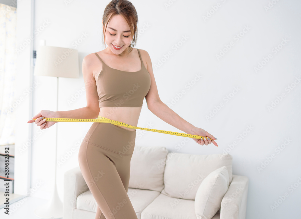 Fototapeta premium young Asian lady in sportswear measuring waist with tape