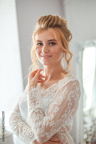 A young beautiful bride with a wedding hairstyle and makeup in the morning in a floor-length white lace tunic near the boudoir table. wedding morning