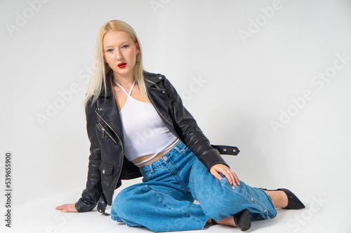 Young beautiful blonde girl in a white top, leather jacket and blue jeans posing on a white background
