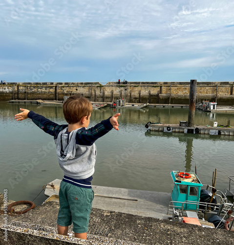 Enfant blond bras levés au bord de l'eau regardant les bateaux et profitant des vacances  photo