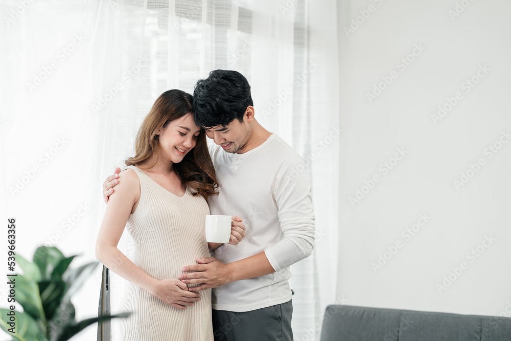 A happy asian couple. A beautiful pregnant wife is holding a cup of milk for the baby with her husband smiling and both of their hands rubbing the wife's tummy in the living room.