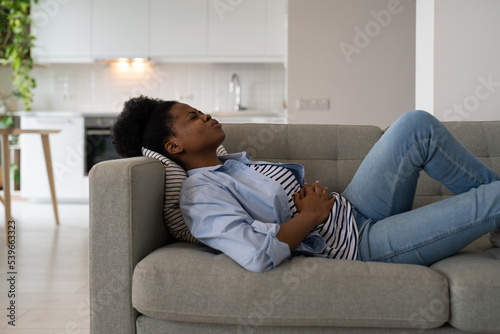 Fotografi Diseased African American woman holding stomach feeling pain and anguish after alimentary poisoning or junk food
