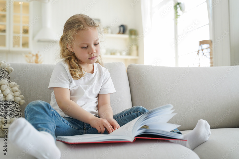 Little elementary child girl reads book, literature bestseller for children, sitting on sofa at home Little elementary child girl reads book, literature bestseller for children, sitting on sofa at home