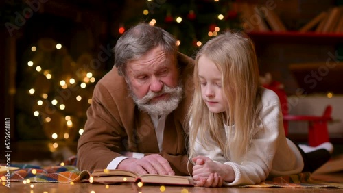 Christmas magic. portrait of joyful caring Caucasian grandfather reading book to granddaughter at home and spending xmas eve near decorated tree. happy holidays concept