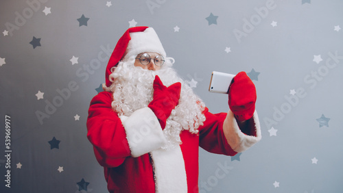 Man in Santa costume is holding smartphone touching screen taking selfie on grey background. Holidays, modern technology and people concept.