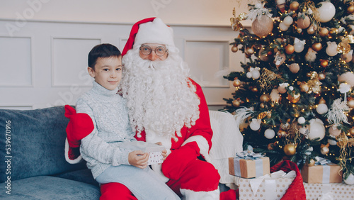 Portrait of adorable child with present and Santa sitting on couch at home looking at camera on New Year's day. People, celebrations and gifts concept.