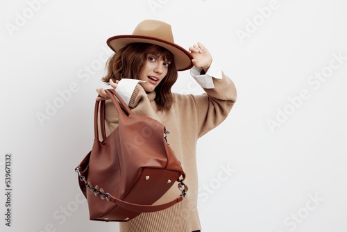 an excited woman poses on a light background in a shirt, sweater and hat, putting her hand on it and throwing a bag over her shoulder