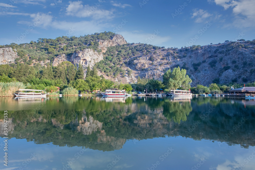 Fototapeta premium The rock-cut temple tombs of the ancient city of Kaunos in Dalyan, Muğla, Turkey. Beautiful view of Dalyan river with reed beds, excursion boats and carved tombs in the background.