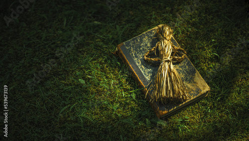 An old book on witchcraft and a voodoo doll lie on the green grass. Items for witchcraft and secret spells. An antique doll made of straw.