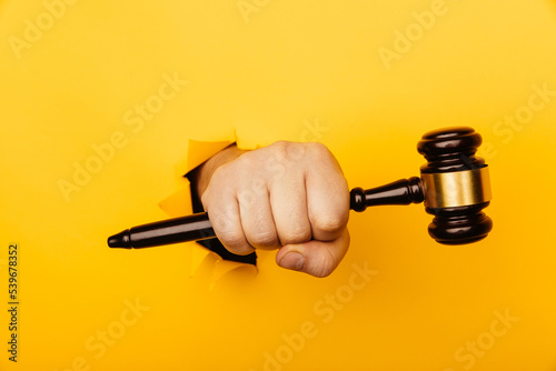 Hand holds a wooden gavel through torn yellow background. Law concept