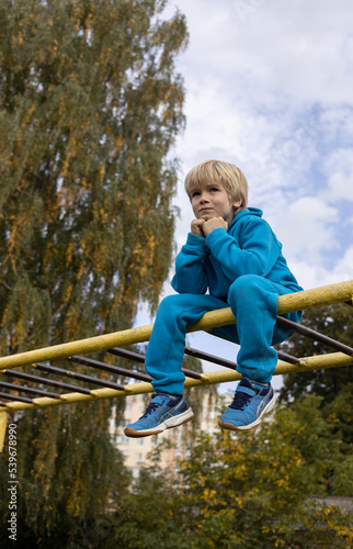 cool schoolboy in a trendy bright blue tracksuit sits on the stairs of the sports ground, view from below. The concept of children's sportswear, active childhood, healthy lifestyle