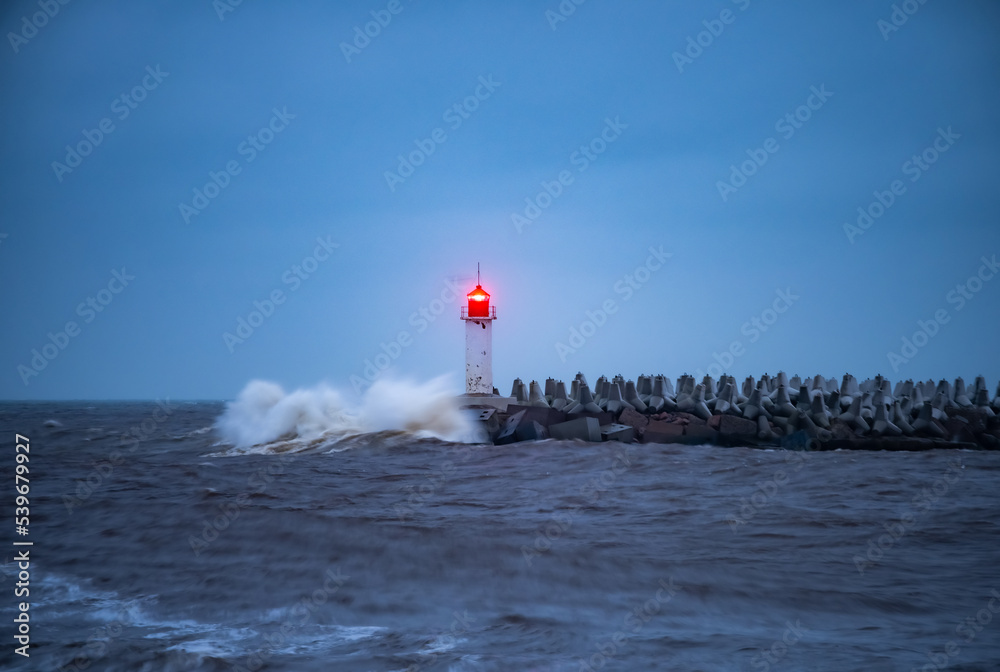 Waves crash against the pier with the lighthouse with red lamp. Baltic sea.