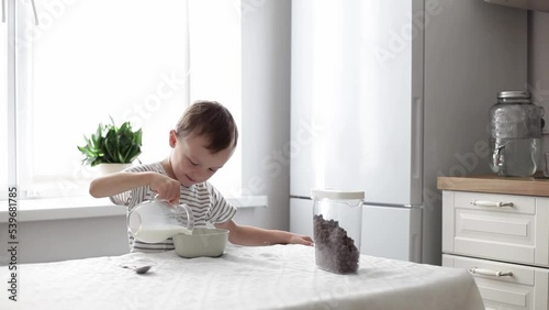 a cute little boy pours milk into a bowl with chocolate flakes. the concept of healthy nutrition.