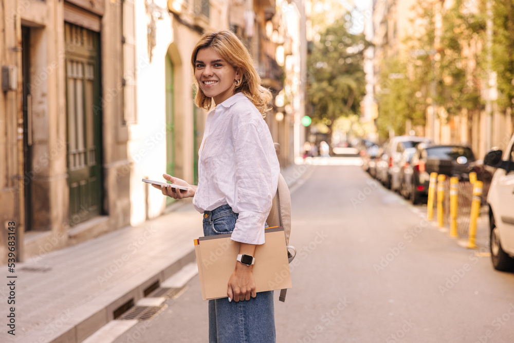 Beautiful european young woman looking at camera, holding phone and notebooks standing on street. Blonde student wear shirt with jeans goes to study in morning. City life concept