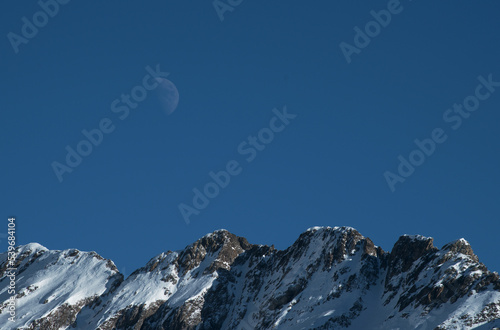 Alpine panorama in the Bergamo Alps, Italy - Winter mountains and moon in the daytime sky