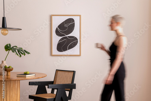 Fotografía Warm and cozy interior of living room space with mock up poster frame, round table, chairs, pedant lamp, rattan chairs and walking young woman