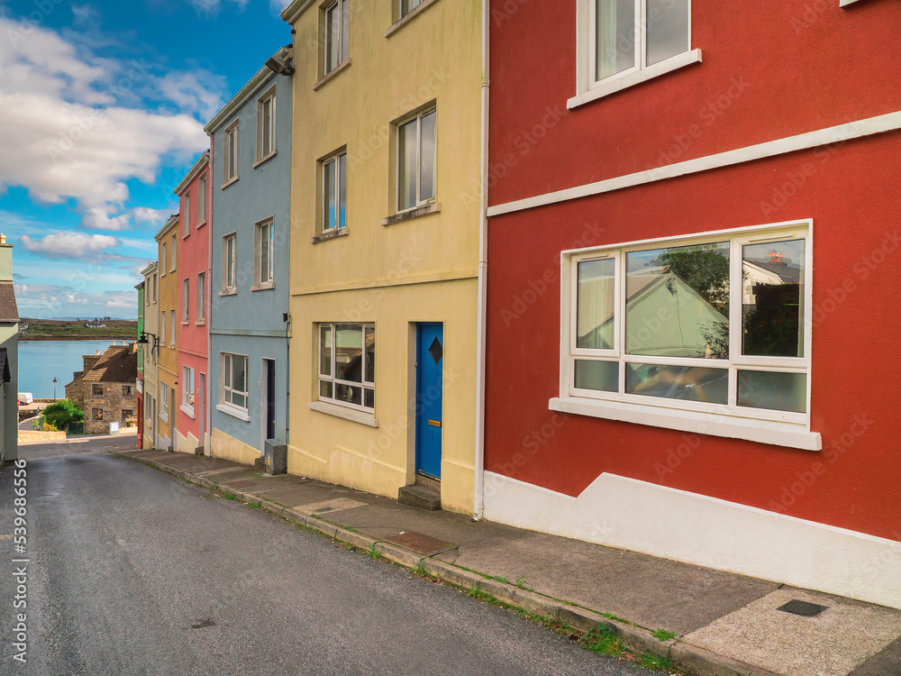 City street with rich saturated color building. Bright painted houses in a town by a small road. Blue cloudy sky. Ocean in the background. West of Ireland.