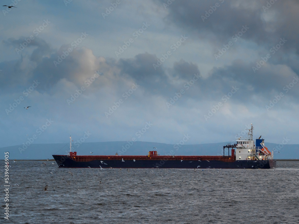 Cargo ship leaving port. Commercial carrier vessel delivering goods and products by water. Import and export concept. Transportation industry. Dramatic cloudy sky.