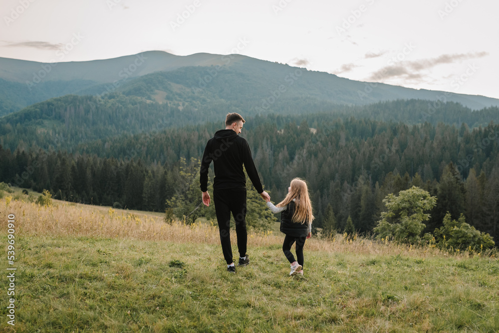 Dad and daughter walk in the mountains enjoy the sunset and look at nature. Back view. The young family spends time together on vacation, outdoors. The concept of family autumn holiday. Father's Day. Dad and daughter walk in the mountains enjoy the sunset and look at nature. Back view. The young family spends time together on vacation, outdoors. The concept of family autumn holiday. Father's Day.