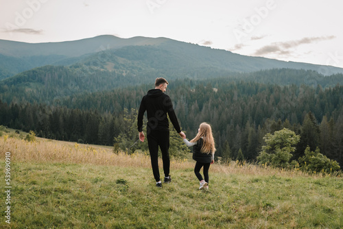 Dad and daughter walk in the mountains enjoy the sunset and look at nature. Back view. The young family spends time together on vacation, outdoors. The concept of family autumn holiday. Father's Day. © Serhii