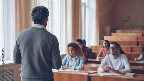 Bearded mature man professor is reading lecture talking and gesturing while students are listening and writing sitting at tables in spacious university classroom.
