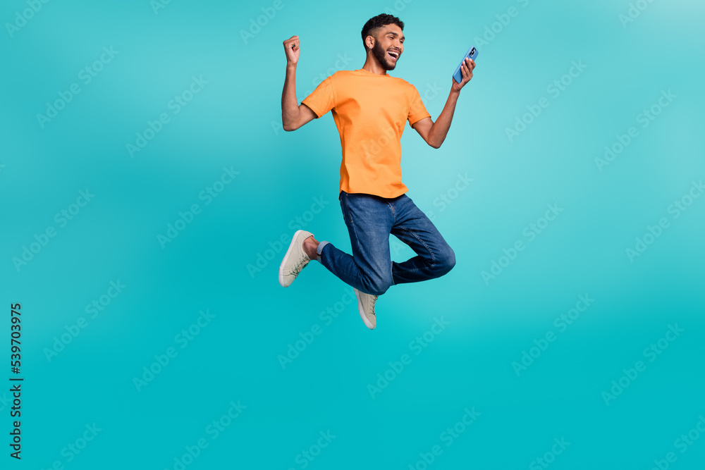 Full length photo of positive lucky guy wear orange t-shirt jumping high rising fist reading modern gadget isolated teal color background