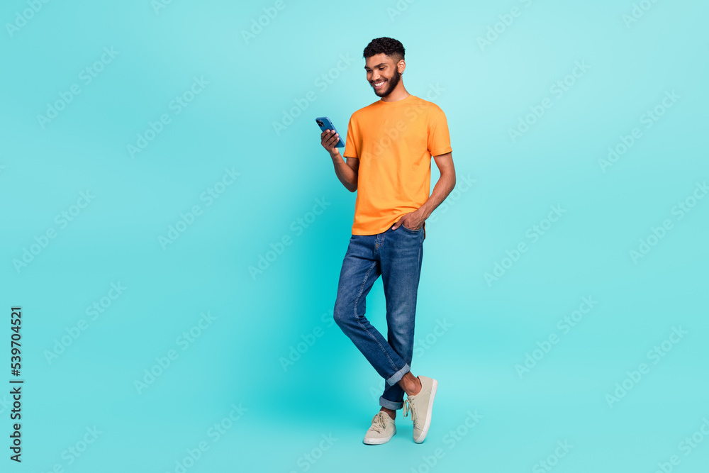 Full length photo of good mood man dressed orange t-shirt typing samsung modern device empty space isolated turquoise color background